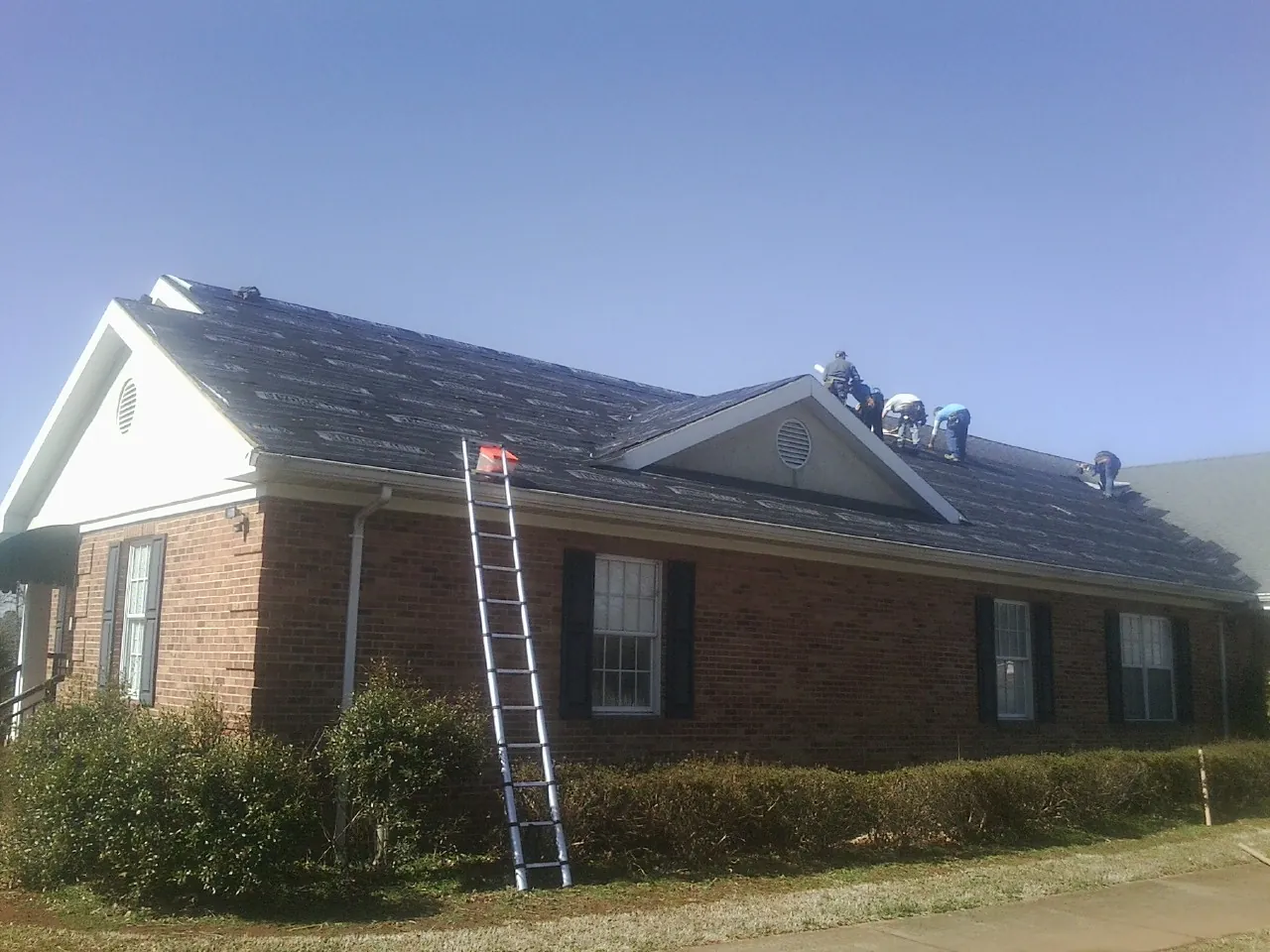Crew tearing shingles off a brick ranch roof with a ladder set — Emergency Roof Repair work by our crew in Oak Island
