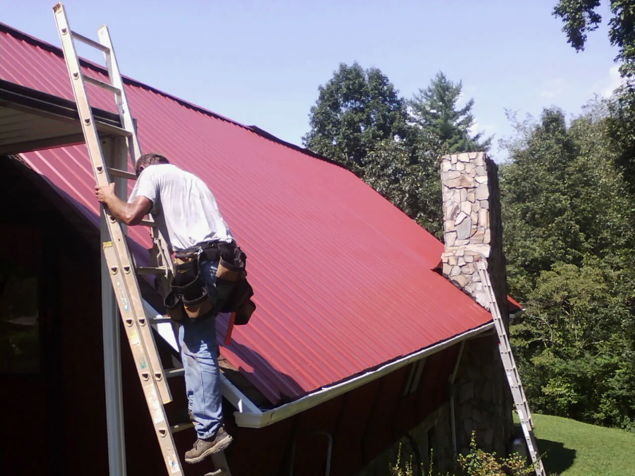 Roofer on a ladder beside a red metal roof and stone chimney — Soffit & Fascia Repair by Roofing Brothers in Oak Island